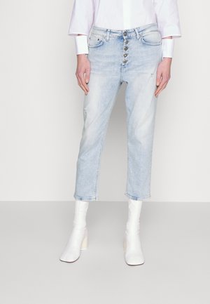 Personne portant un jean court bleu clair à fermeture par boutons, des bottines blanches à talons et une chemise blanche à manches longues sur un fond uni.