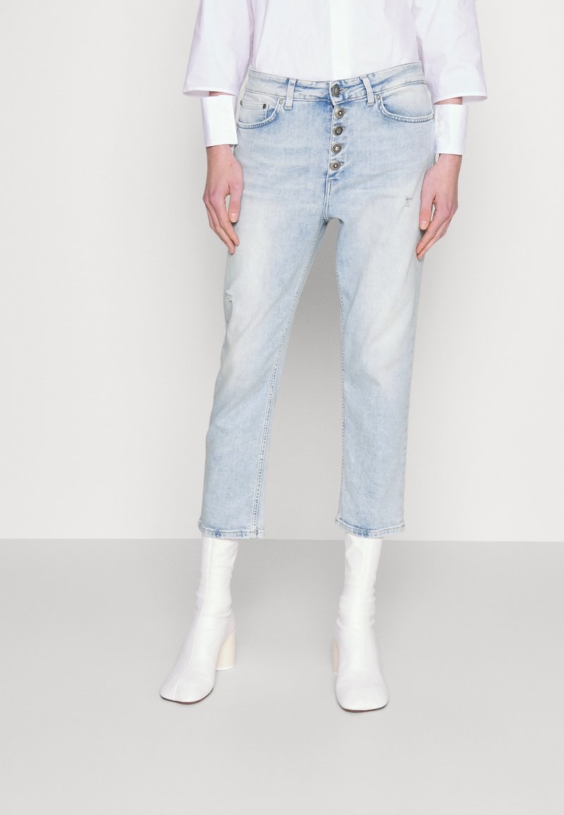 Personne portant un jean court bleu clair à fermeture par boutons, des bottines blanches à talons et une chemise blanche à manches longues sur un fond uni.