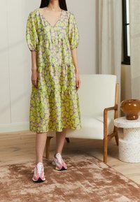 Zelené a fialové vzorované vrstvené šaty s výstřihem do V a balónovými rukávy, nošené s růžovými teniskami na dřevěné podlaze blízko židle.