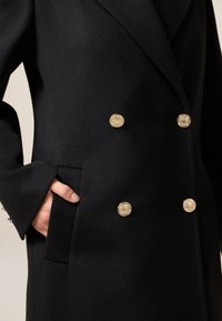 Cappotto di lana nero con design doppiopetto, bottoni dorati, maniche a campana e tasca laterale. Tessuto con texture visibile.