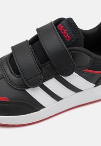 Zapatilla deportiva sintética negra con diseño de tres franjas blancas, tiras de Velcro, acentos rojos y un upper texturizado.