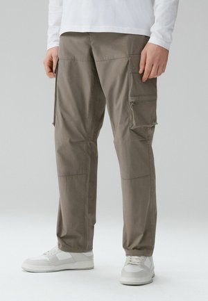 Man wearing olive cargo pants with multiple pockets and white sneakers, standing against a plain light background.