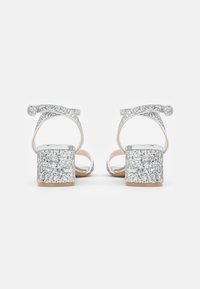 Sandali con tacco a blocco in argento glitterato con cinturino alla caviglia. Il cinturino presenta un design incrociato, che aggiunge texture e brillantezza.
