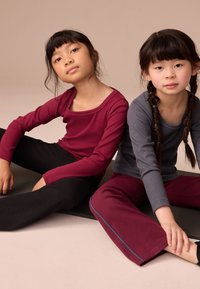 Dve dekleti sedita na preprogi. Ena nosi bordo majico z dolgimi rokavi s širokim izrezom in rebraste črne hlače. Druga nosi sivo majico in bordo hlače.