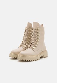 Bottes chelsea beige à lacets en cuir avec accents dorés et semelle épaisse. Surface texturée et cheville rembourrée pour le confort.