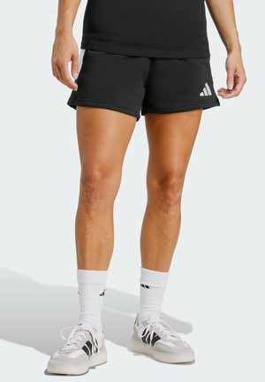 Osoba nosící černé šortky Adidas, bílé ponožky Adidas a bílé tenisky s černými pruhy, stojící před bílým pozadím.