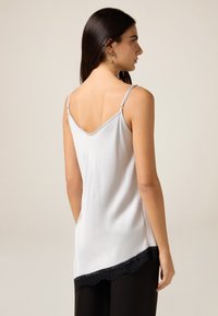Lysegrå ærmeløs top med v-udskæring, der har sort blonderkant ved kanten. Glat tekstur og tynde justerbare stropper.