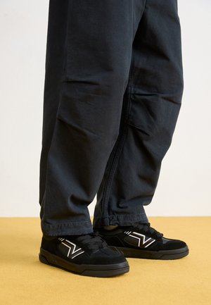 Black sneakers made of suede and synthetic materials, featuring white accents and a flat rubber sole, paired with dark baggy pants.