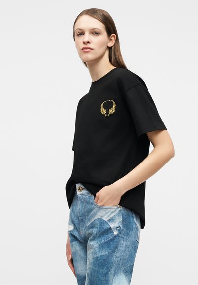 Γυναίκα που φοράει μαύρο μπλουζάκι με μικρό χρυσό σχέδιο ιππόκαμπου στο στήθος, συνδυασμένο με ανοιχτόχρωμο τζιν, με το χέρι στην τσέπη.