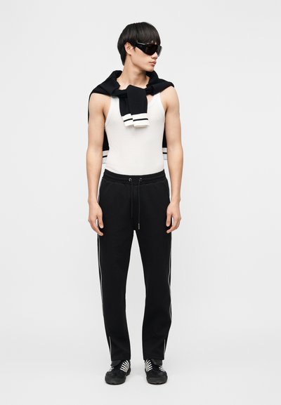 Νεαρός άνδρας που φοράει μαύρα γυαλιά ηλίου, λευκό αμάνικο πουκάμισο, μαύρο παντελόνι με ρίγες στα πλάγια, μαύρο πουλόβερ που κρέμεται από τους ώμους και μαύρα αθλητικά παπούτσια.