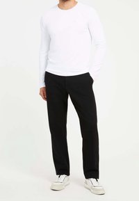 Vit långärmad skjorta med rundad halsringning, ihop med svarta byxor. Modellen bär vita sneakers; slät textur och minimalistisk design.