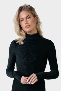 Schwarzes, geripptes, langärmliges Rollkragenoberteil mit figurbetontem Design. Das Model zeigt Hände, die mit mehreren Ringen geschmückt sind, vor einem neutralen Hintergrund.