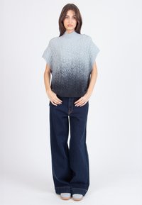 Žena stoji u širokim tamnim trapericama i sivoj ombre pletenoj bezrukavnoj rolci nasuprot blijede bijele pozadine.