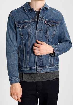 Person iført en blå denimjakke over en mørkegrå skjorte, med en hånd, der justerer jakken nær brystet.