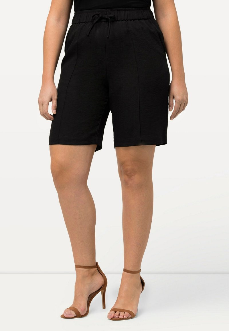 Femme portant un short noir jusqu'aux genoux avec une taille à cordon et des sandales à talons hauts marron, debout devant un fond blanc.