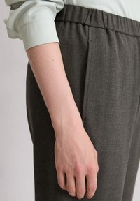 Pantalons gris texturés avec une taille élastique et des poches latérales, présentés à côté d'une manche de blouse de couleur neutre près d'une main.