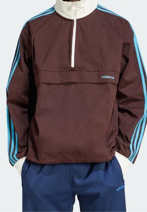 Man draagt een bruine Adidas trui met lichtblauwe strepen en een witte rits, gecombineerd met blauwe Adidas korte broek.