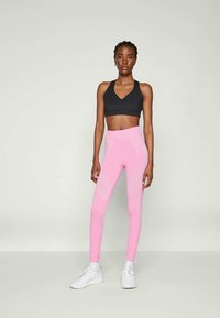 Reggiseno sportivo nero e leggings rosa con texture a coste diagonali, abbinati a sneakers bianche. Sfondo semplice.