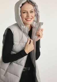 Jasnoszara kamizelka puchowa z kapturem, przednim zamkiem błyskawicznym, elastycznymi sznurkami oraz bocznymi kieszeniami. Nosi się ją na czarnym topie z długim rękawem.