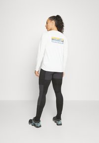 Wit T-shirt met lange mouwen met een veelkleurig Patagonia-logo op de achterkant, gecombineerd met zwarte leggings met grijze accentpanelen en sneakers.