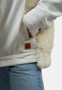 Veste en denim blanc avec doublure en fausse fourrure crème, ornée d'un patch en cuir étiqueté "R.D.K.S." et de poignets distressed, associée à un jean bleu clair.