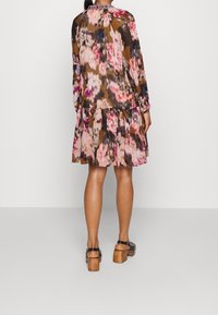 Kukkapainotteinen mekko, jossa pitkät hihat, kerroksittainen helma ja ruskea pohjaväri. Pyöreä pääntie ja teksturoidun näköinen kangas. Ruskeat korolliset sandaalit.