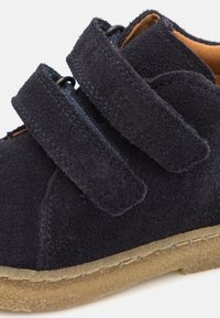 Zapato de ante azul marino con dos correas ajustables de velcro, puntera redonda y suela de goma beige. Contiene detalles de costura visibles.