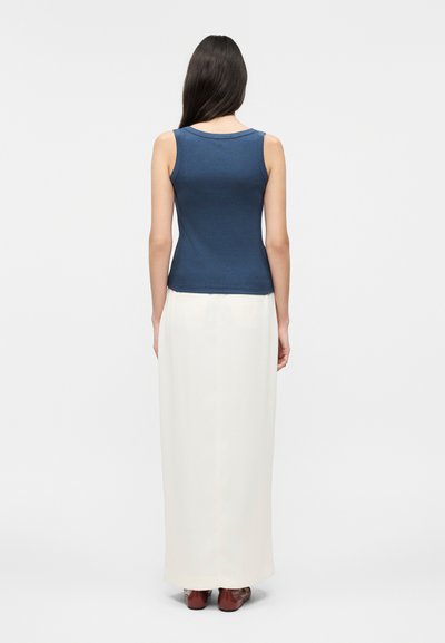 Γυναίκα με μακριά σκούρα μαλλιά, που φοράει μπλουζάκι χωρίς μανίκια σε ναυτικό μπλε χρώμα και μακριά λευκή φούστα, στέκεται με την πλάτη προς το μέρος του θεατή σε ένα απλό φωτεινό φόντο.