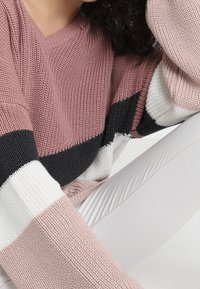 Gestrickter Pullover mit horizontalen Streifen in Pink, Schwarz und Weiß. Lockerer Schnitt mit weiten Ärmeln und strukturierter Stoff. Hosen in neutralen Tönen sichtbar.