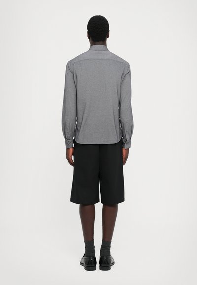 Un uomo di spalle che indossa una camicia grigia a maniche lunghe, pantaloni corti neri fino al ginocchio, calze scure e scarpe eleganti nere, su uno sfondo chiaro.