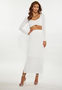Witte lange mouwen crop top met geplooide details, gecombineerd met een witte maxi rok met een elastische tailleband en een split aan de zijkant, aangevuld met zilveren hakken.