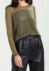 Top verde oliva con maniche lunghe trasparenti e corpino solide, abbinato a pantaloni in pelle nera a vita alta con dettagli plissettati.