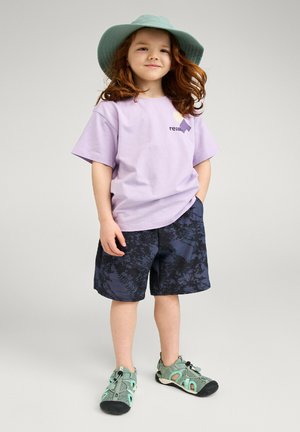 Dziecko z długimi czerwonymi włosami nosi zielony kapelusz typu bucket, jasnofioletową koszulkę, ciemne spodenki w wzory oraz zielone sportowe sandały, stoi z rękami w kieszeniach.