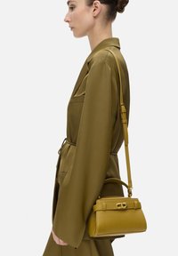 Mosterdgele handtas met een gestructureerde vorm, voorzien van een handvat aan de bovenkant en een verstelbare schouderband. Gladde lederen materiaal met metallic accenten.