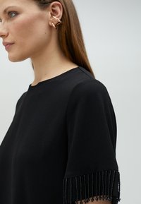 Crna bluzica kratkih rukava s ukrasnim rese na манжетама. Glatka tkanina s klasičnim okruglim izrezom.