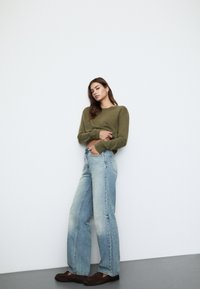 Olivno zeleni pulover z dolgimi rokavi, svetlo modri široki kavbojki in temno rjavo obutev. Gladka tekstura in sproščen kroj.