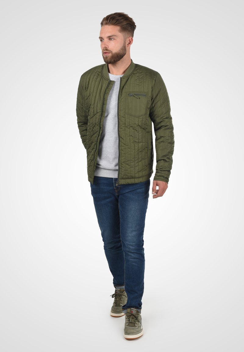 Groene gewatteerde bomberjack met een ritszak, gedragen over een grijze trui. Gecombineerd met blauwe jeans en olijfgroene sneakers.