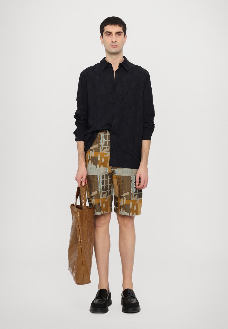 Sort skjorte med knapper og tekstureret mønster, mønstrede brune shorts, brun shoppingtaske og sorte loafers. Model står foran en ensfarvet baggrund.
