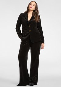 Completo in velluto nero con blazer aderente caratterizzato da due bottoni dorati e revers classici; pantaloni a gamba larga con una texture liscia.