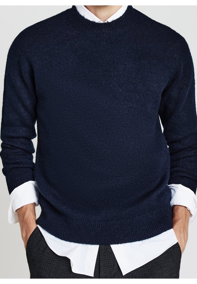 Homme portant un pull en maille bleu foncé par-dessus une chemise blanche boutonnée, les mains dans les poches, montré du cou jusqu'aux hanches.