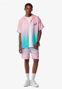 Ensemble chemise à manches courtes à boutons et short, dégradé du rose au bleu sarcelle, texture à motifs, avec logo de palmier, accompagné de baskets blanches.