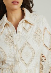 Camicia bianca con bottoni, caratterizzata da un intricato ricamo in pizzo beige con motivi floreali e a diamante. Tessuto di cotone morbido, colletto strutturato.