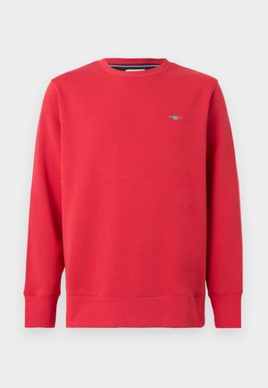 Piros pulóver kerek nyakkivágással, hosszú ujjakkal és bordás mandzsettákkal. Kis logó található a mellrészen. Puha anyagból készült.