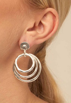 Primer plano de una oreja rubia que lleva un pendiente plateado con tres aros circulares irregulares enlazados colgando de un botón.