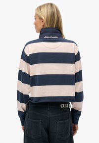 Marineblauw en lichtroze gestreepte polo met lange mouwen, een hoge kraag, een cropped ontwerp en geborduurde "Athletic Essentials"-detail op de achterkant.