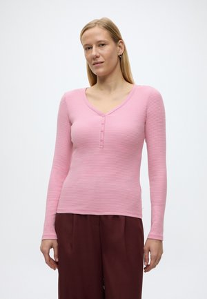 Frau mit glattem blondem Haar trägt ein rosa, langärmeliges, geknöpftes Oberteil und bordeauxrote Hose, steht vor einem schlichten weißen Hintergrund.