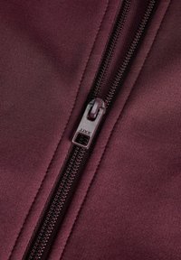 Gros plan d'une fermeture éclair violet foncé sur un tissu assorti avec des surpiqûres visibles et un curseur de fermeture portant l’étiquette JJXX.