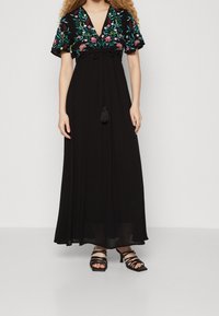 Schwarzes Maxikleid mit kurzen, bestickten Blumensäulen und einem gebundenen Taillenband. Verfügt über einen V-Ausschnitt und einen fließenden, transparenten Rock. Schwarze, strapazierfähige Absätze.