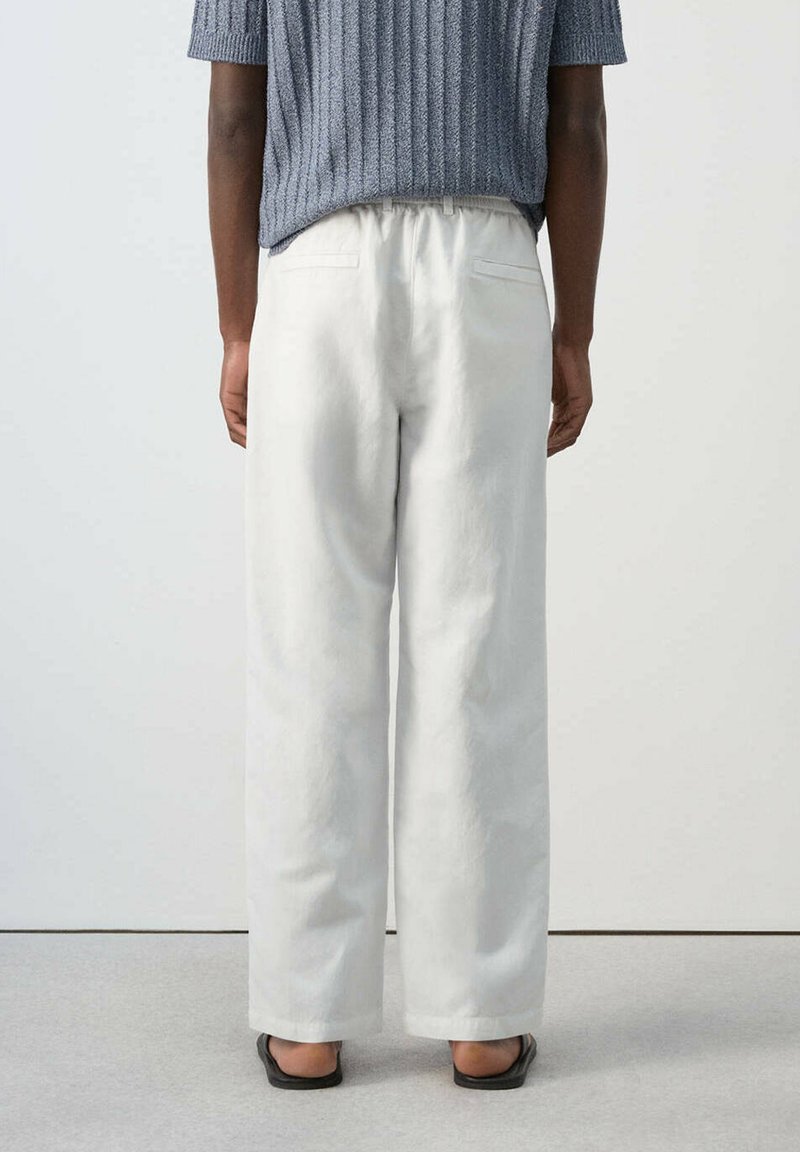 Personne portant un pantalon blanc ample, un haut en maille gris à manches courtes et des chaussures plates noires ouvertes au talon, debout devant un fond clair uni.