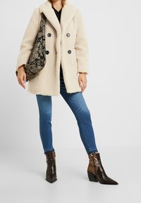 Mujer vistiendo un abrigo de peluche beige, jeans ajustados azules, botas de tobillo de serpiente marrón y llevando un bolso de hombro de patrón negro y beige.